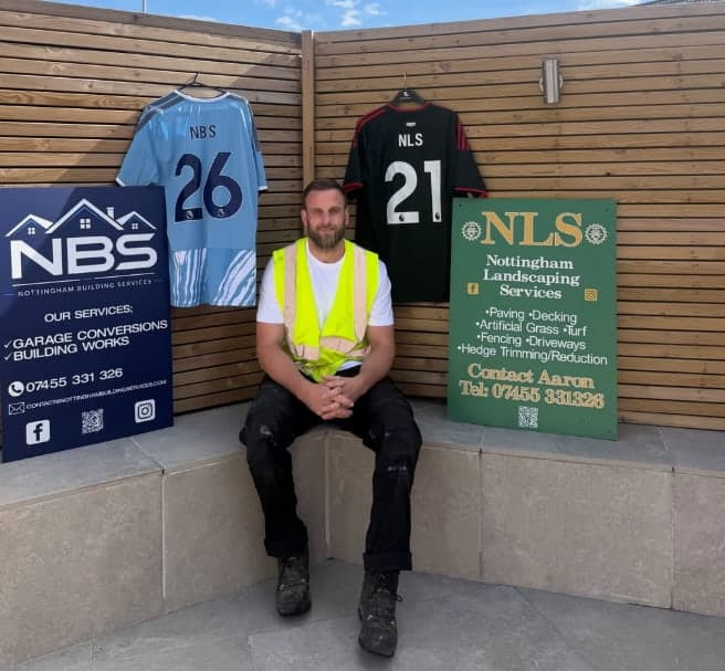 Aaron Brereton sitting between an NBS board and an NLS board, with an NBS shirt and an NLS shirt hung on the fence behind him